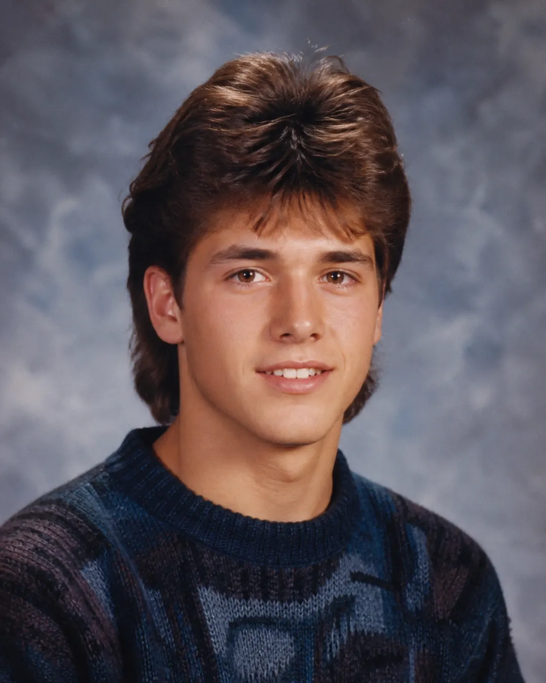 A fictional young adult in a believable 1980s school-yearbook portrait with a patterned sweater and mottled blue-gray backdrop.